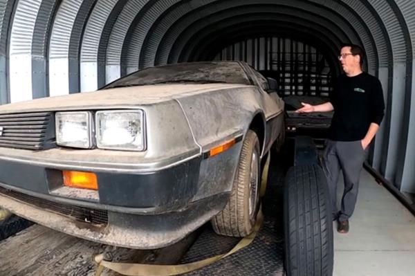 A 1981 Delorian sits on a trailer with a man standing next to it.