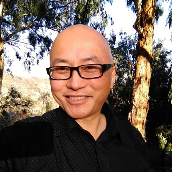Iwamasa wears black rimmed glasses and a black collared shirt and smiles for a selfie in front of trees
