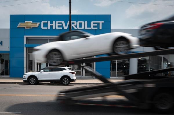 A car hauler passing a Chevrolet dealership in Chicago, representing the halted workflow due to a cyber attack on auto dealership software systems