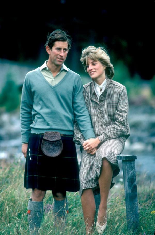 A photo of Charles, in a blue sweater and kilt, and Diana, in a white blouse, plaid overcoat and beige shoes, holding hands in a field.