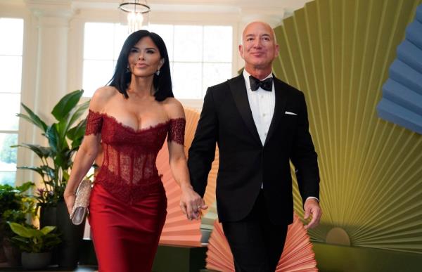 Jeff Bezos in a tuxedo and Lauren Sanchez in a plunging red dress walking hand in hand.