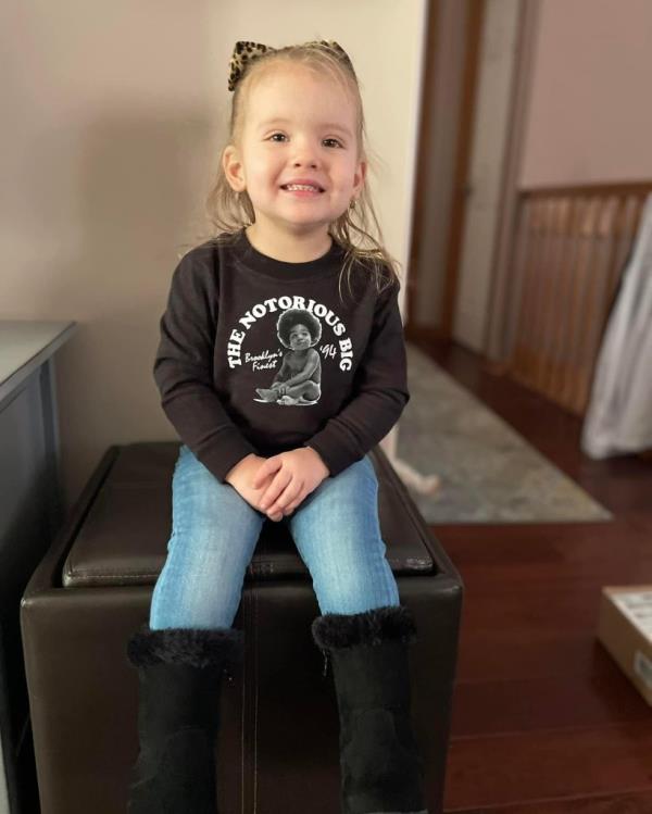 A little girl smiling in a bedroom wearing a Notorious B.I.G. shirt.