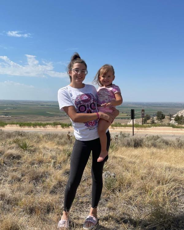 A young woman holds a little girl with a prairie in the background.