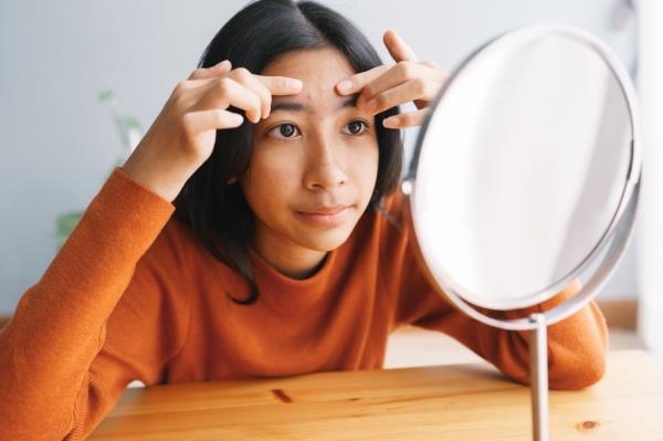 Young Asian woman looking in mirror while touching her acne skin at home