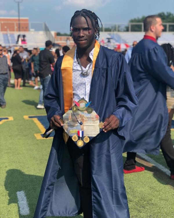 Mario Heslop in a graduation gown holds a decorated cap showing his athletic career with other graduates in the background on a school field. 