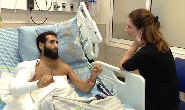 Raz Peri sits in his hospital bed with his arm in a cast and speaks to a woman with her elbow leaning on the bed's side.