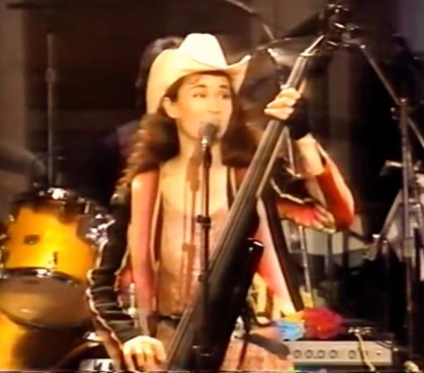 Laura Lynch seen in early performance playing bass and singing on stage wearing cowboy hat.