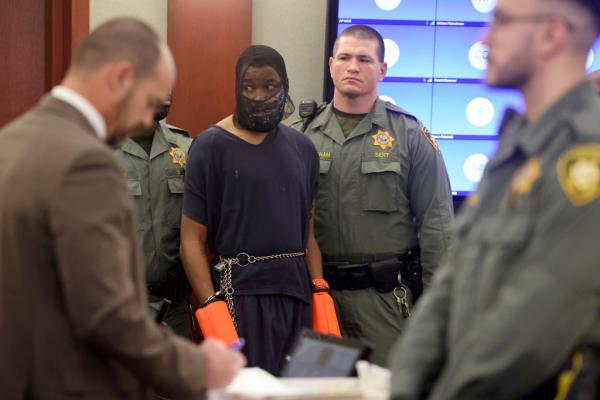 A man wearing a spit mask surrounded by police officers in a courtroom.