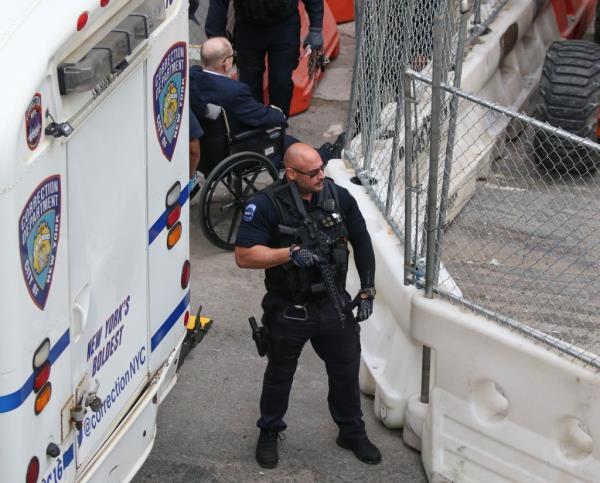 Harvey Weinstein in a suit and tie, sitting in a wheelchair, being escorted by armed police officers out of Bellevue Hospital in New York