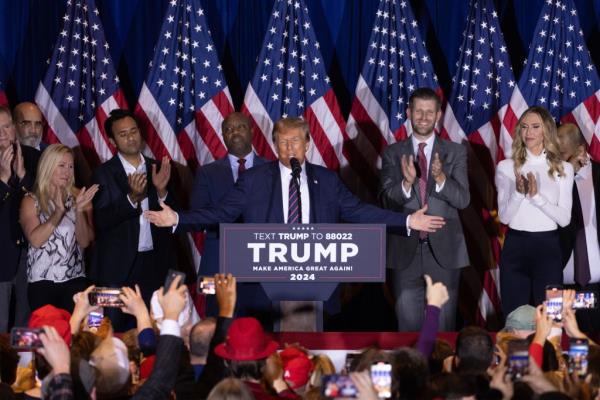 Do<em></em>nald Trump addresses fans at a rally. He stands on stage and speaks into a microphone while holding his arms out wide as his family members and supporters who are standing around him applaud along with the crowd