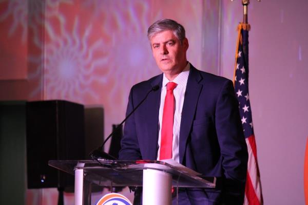 Fulton County Commissio<em></em>ner Bob Ellis wears a navy suit and red tie as he speaks into a microphone fixed to a lectern and with a USA flag behind him