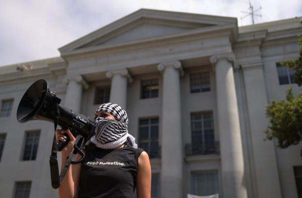 A pro-Palestinian protester WITH THEIR FACE COVERED BY A KEFFIYEH uses a bullhorn.