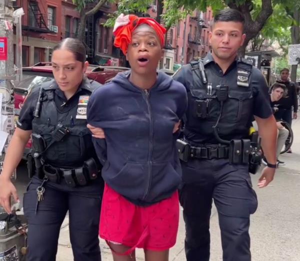 Two police officers grab the arms of a cuffed woman in a sweatshirt, red shorts and red durag.