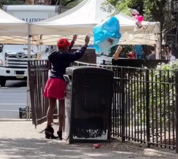 Woman in red shorts throws a blue trash bag over a fence into an area with plants in the park. 