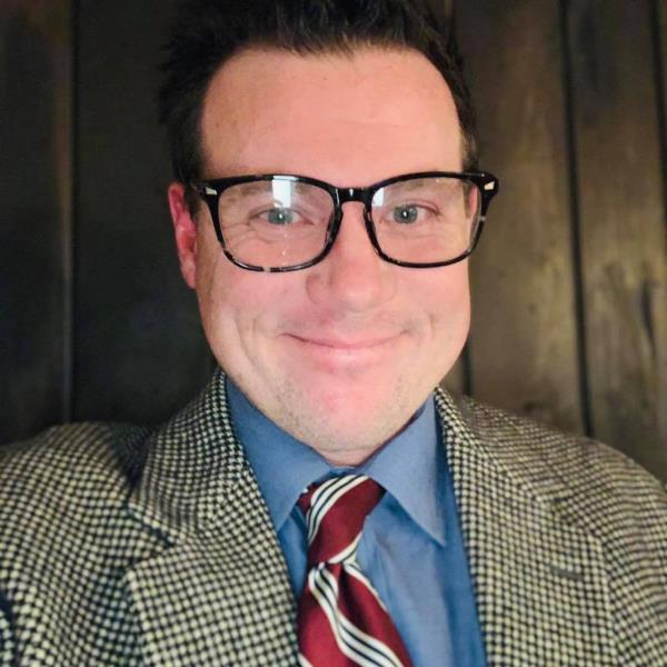 A man with black glasses, a black-and-white jacket and a blue button-down with a red, white and black striped tie.