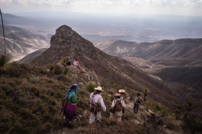 Wixarika Indigenous people walk over a mountain with views of the desert plain below