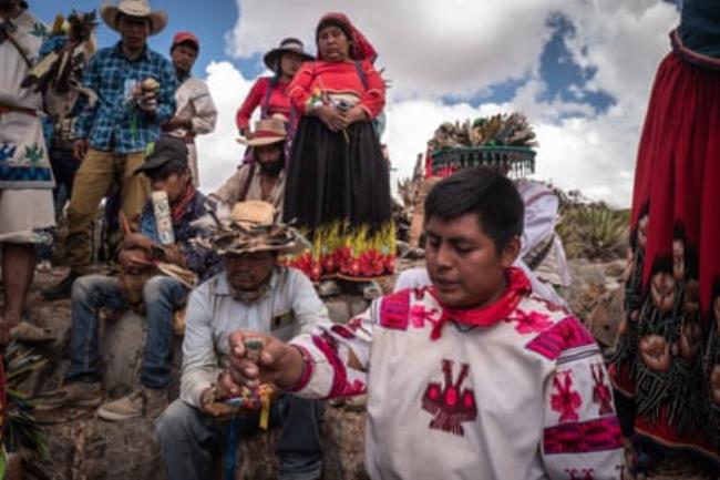 An Indigenous man with closed eyes, wearing a white shirt with traditio<em></em>nal pink embroidery on it, holds out a piece of peyote, as a group of Indigenous people watch him