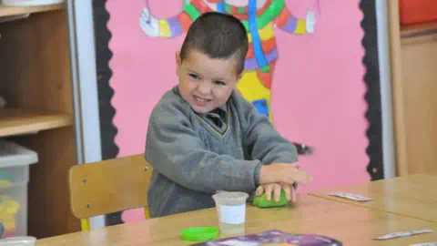 Newspics A young child in a school uniform stands next to a table. He is playing with a green item that looks like clay.