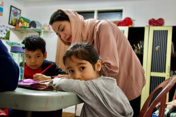 An Afghan women teaching refugee children in Indonesia. One child has turned to look at the camera with their elbows over their books. The teacher is wearing a pale pink outfit with a headscarf tied loosely around her dark hair.